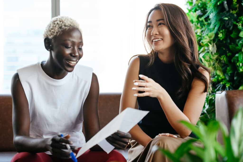 Two professional women communicating over a piece of paper. they are smiling and working collaborating in a casual office environment