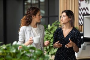 Two female colleagues walk along in conversation in the office, one holding a coffee cup