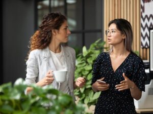 Two female colleagues walk along in conversation in the office, one holding a coffee cup