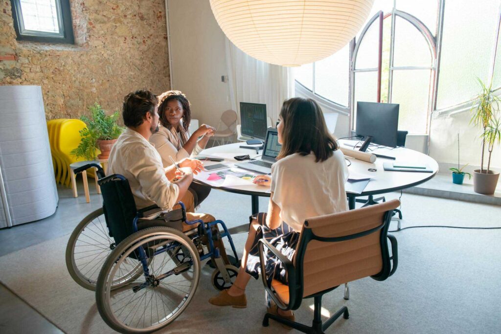 A trio of work colleagues sit at a round table having a meeting, one in a wheelchair and the other two sat beside him